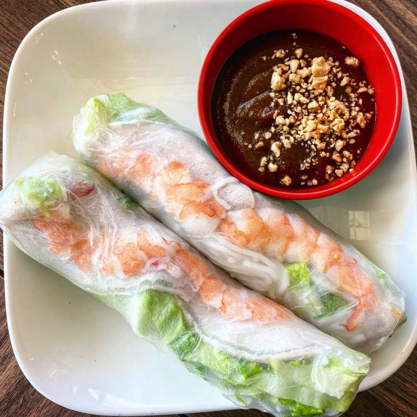 Two fresh Vietnamese spring rolls with shrimp and lettuce, served with a red bowl of peanut sauce.